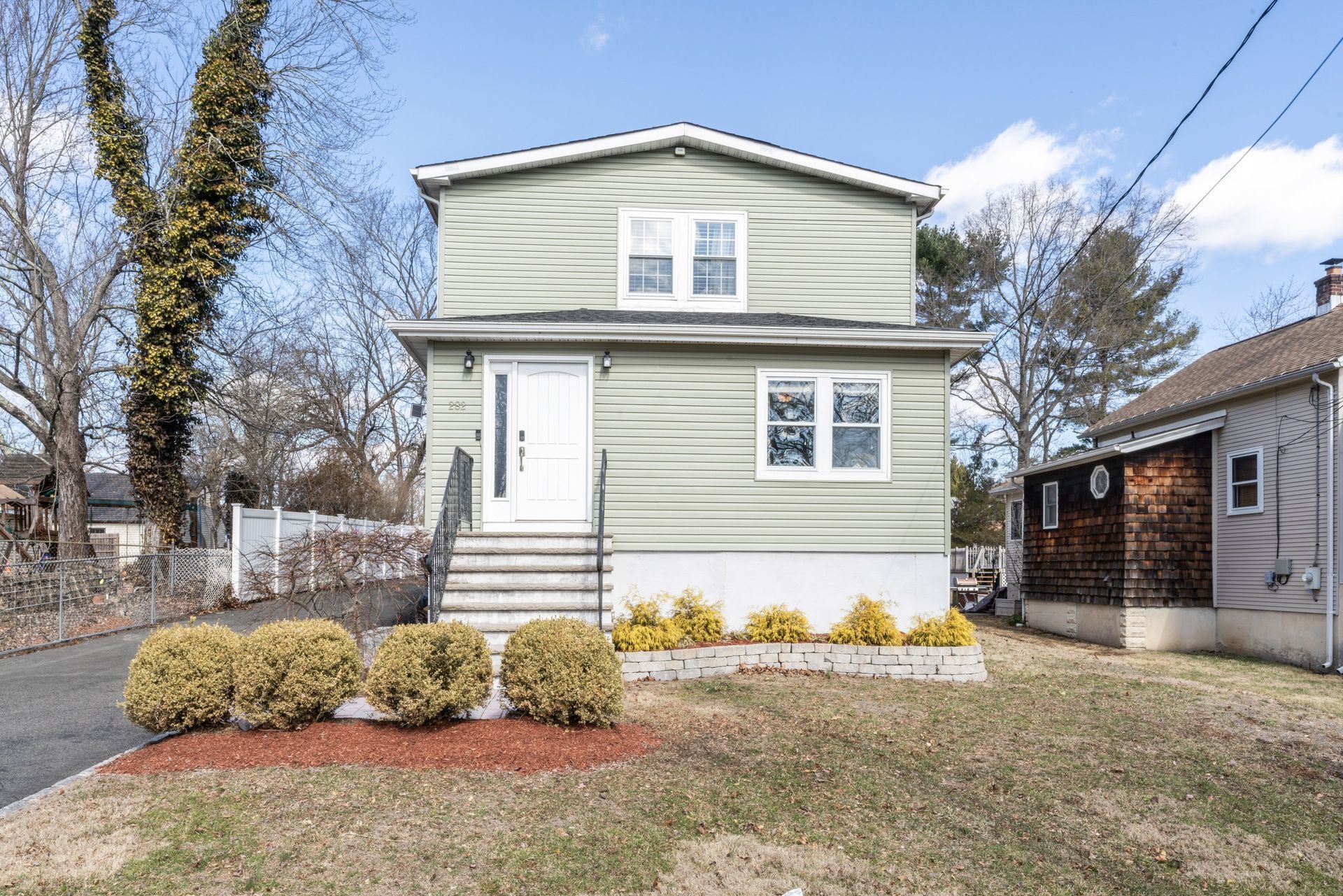 Two-story house with green siding, white door, and steps leading to entrance, set in a yard with shrubs.