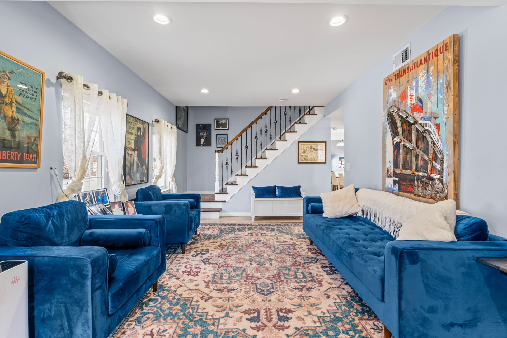 Living room with blue velvet furniture, colorful rug, and artwork. Staircase in background.