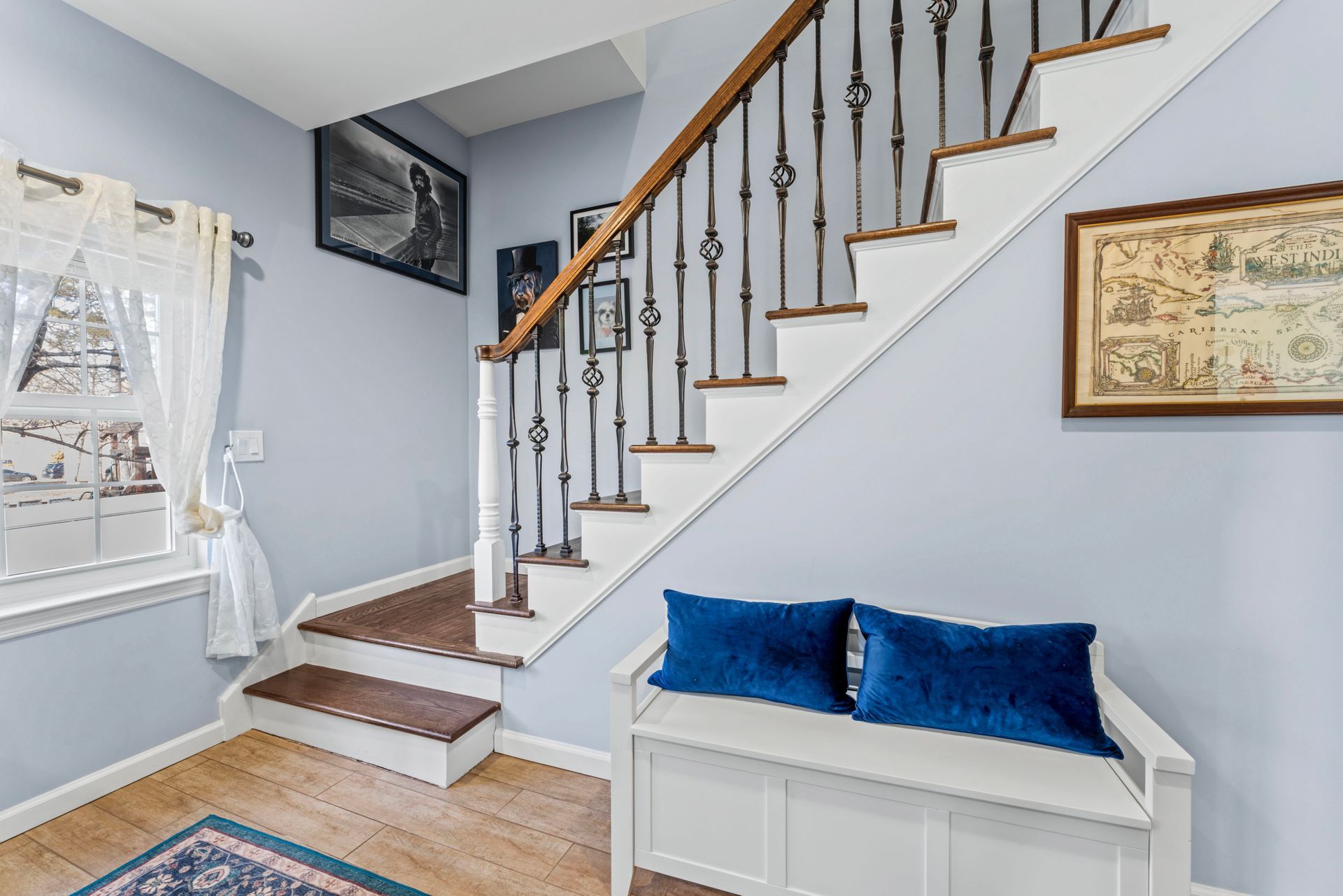 Entryway with staircase, white bench with blue pillows, and framed artwork on the walls.