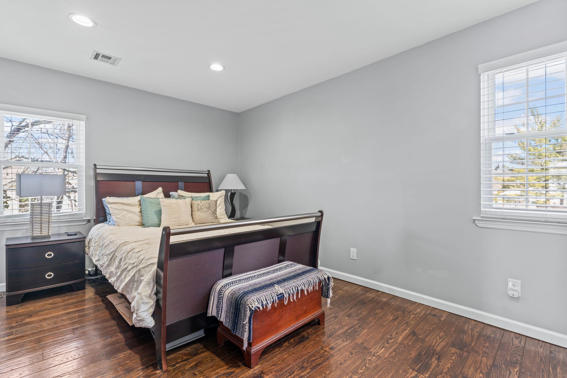 Bedroom with a bed, nightstand, chest, two windows, and dark wood floors. Gray walls.