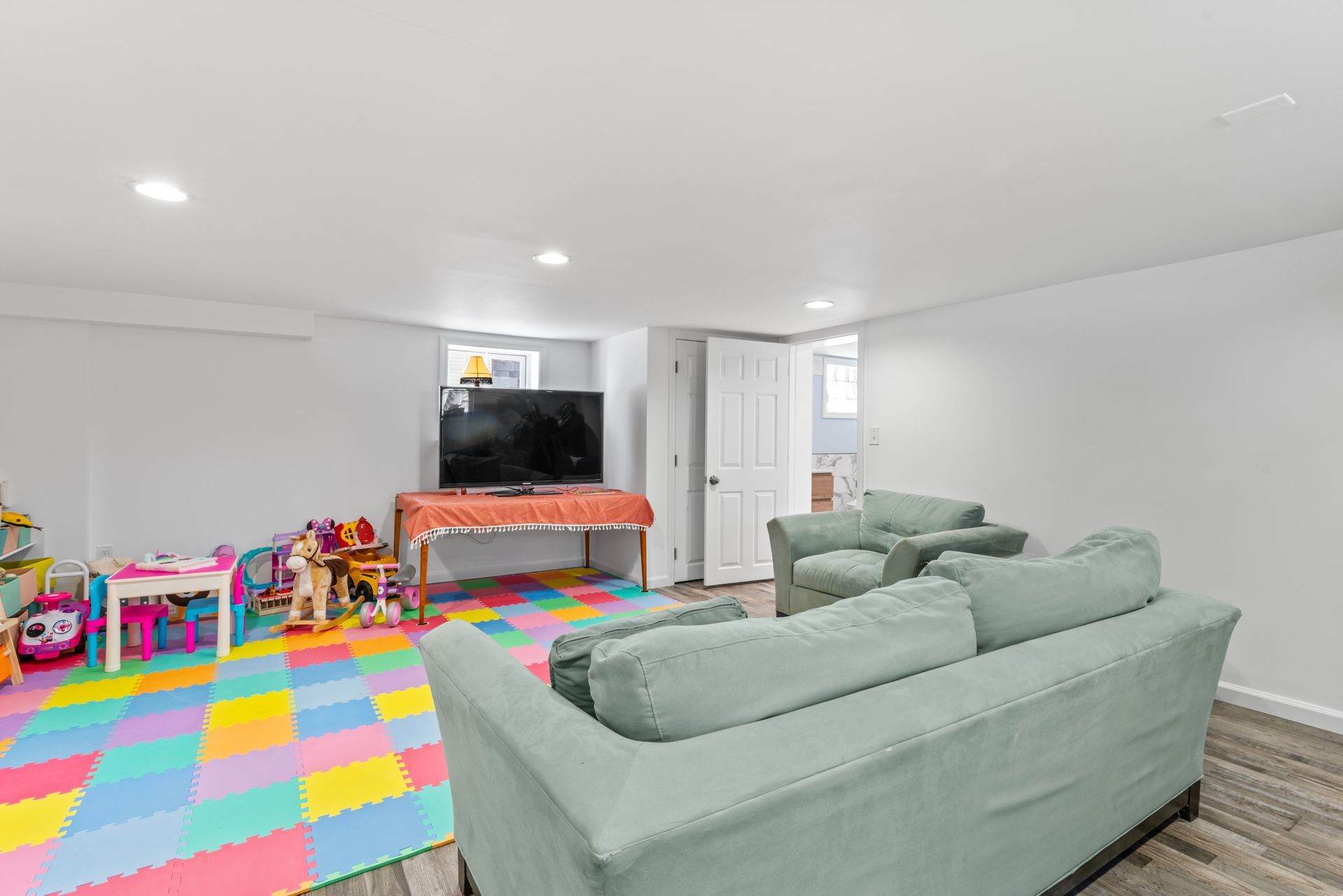 Living room with couches, a TV, toys on colorful floor mats, and a door.