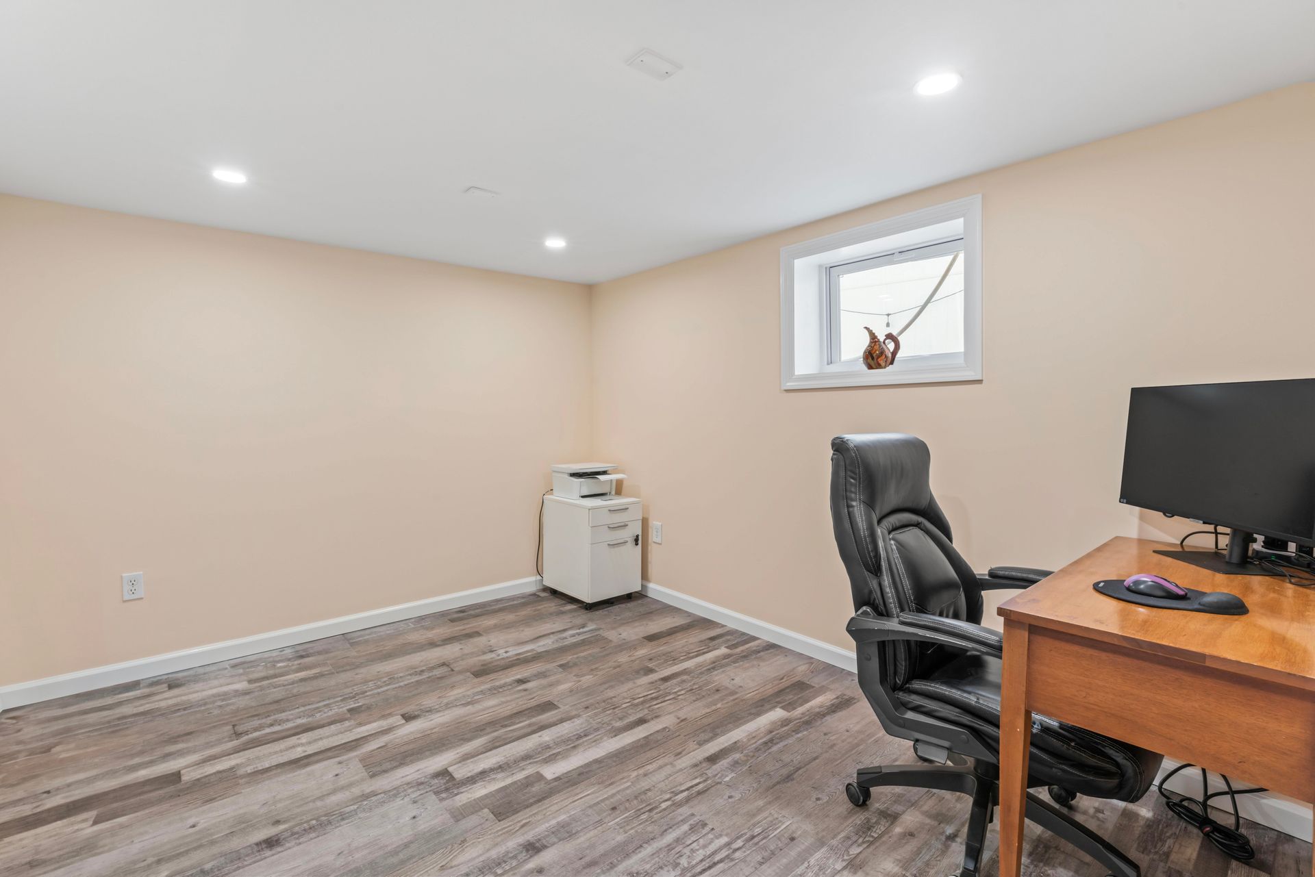 Home office with desk, chair, printer, and window. Beige walls, wood-look floor, and bright lighting.