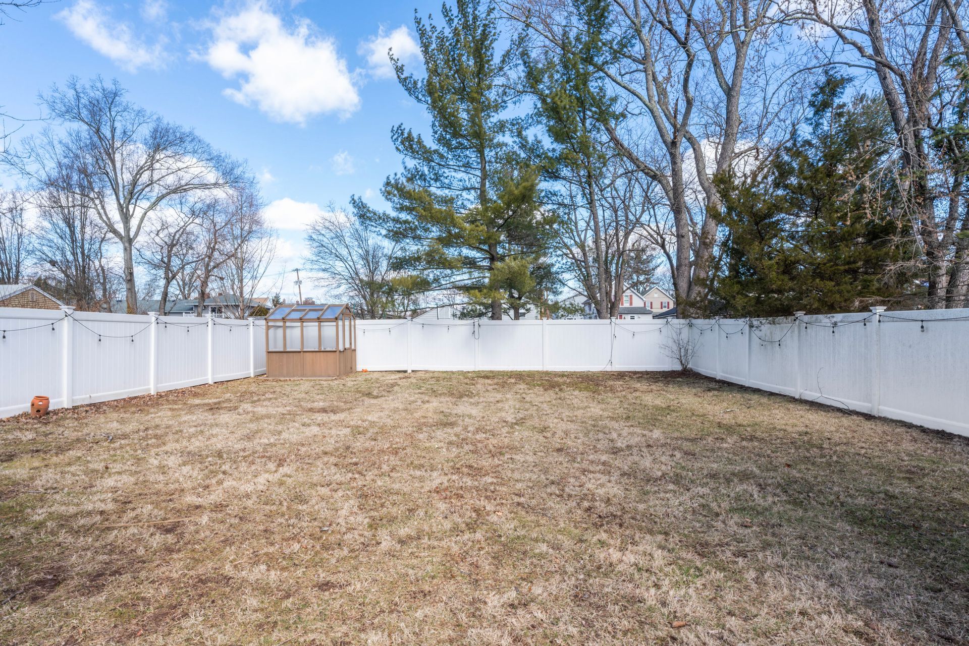 Backyard enclosed by a white fence, dry grass, with a small shed and trees against a blue sky.