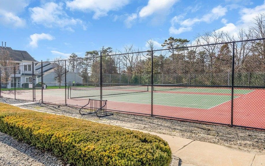Tennis court behind a chain-link fence, with a red and green surface, trees in the background, and a blue sky with clouds.