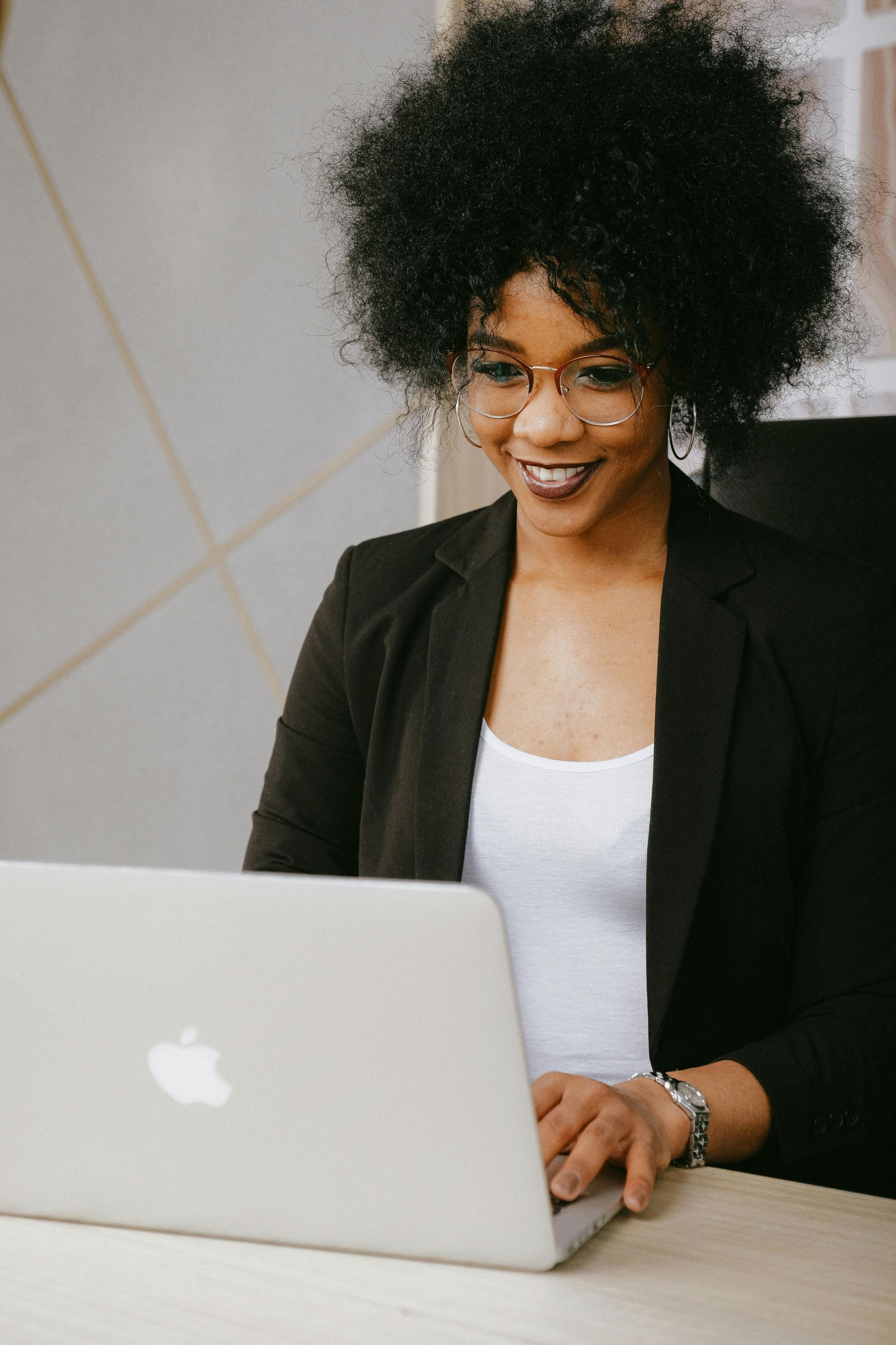Woman smiling while working on a laptop, wearing a blazer, white shirt, and glasses.
