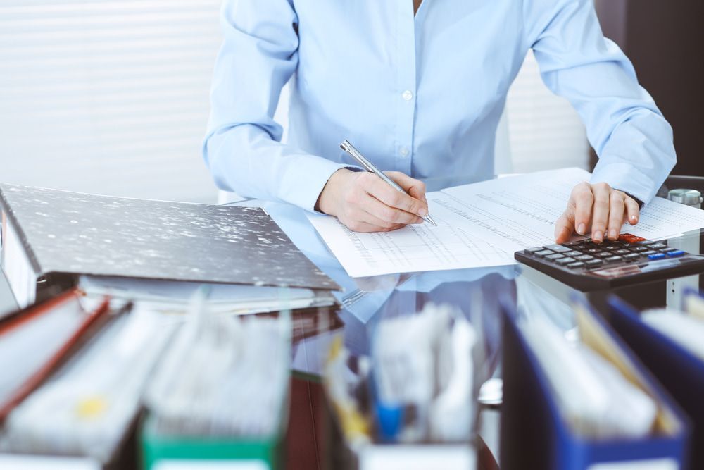 Person writing on paper with a pen at a desk, using a calculator surrounded by files. — Sunshine Office Solutions in Buderim, QLD