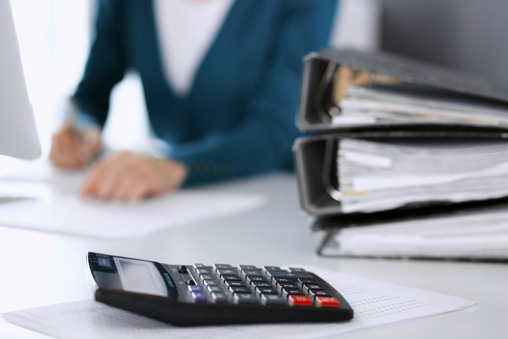 Calculator on desk with stack of binders and person in background. — Sunshine Office Solutions in Buderim, QLD