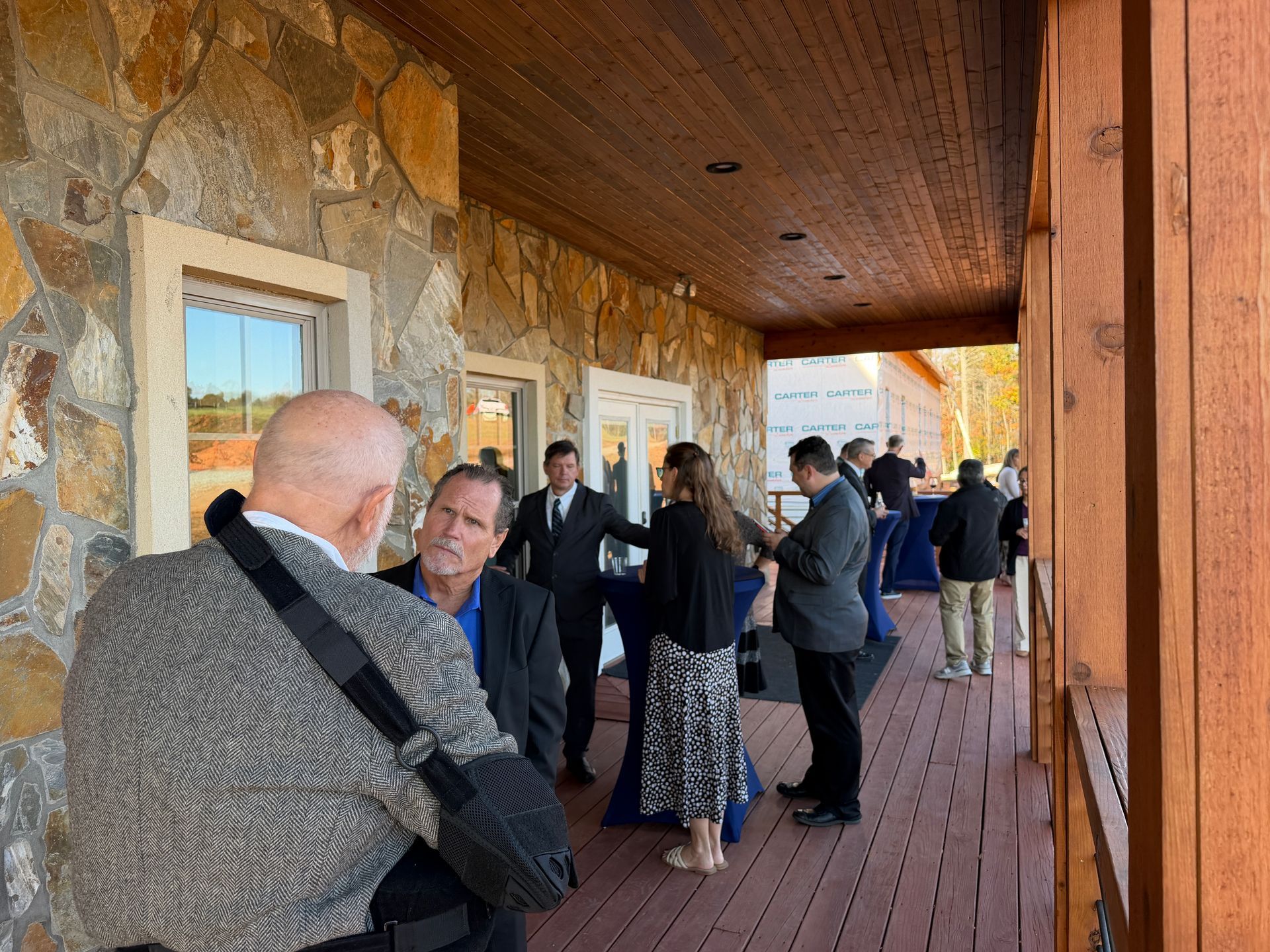 People gathered on a covered stone porch, chatting near windows and a wooden ceiling.