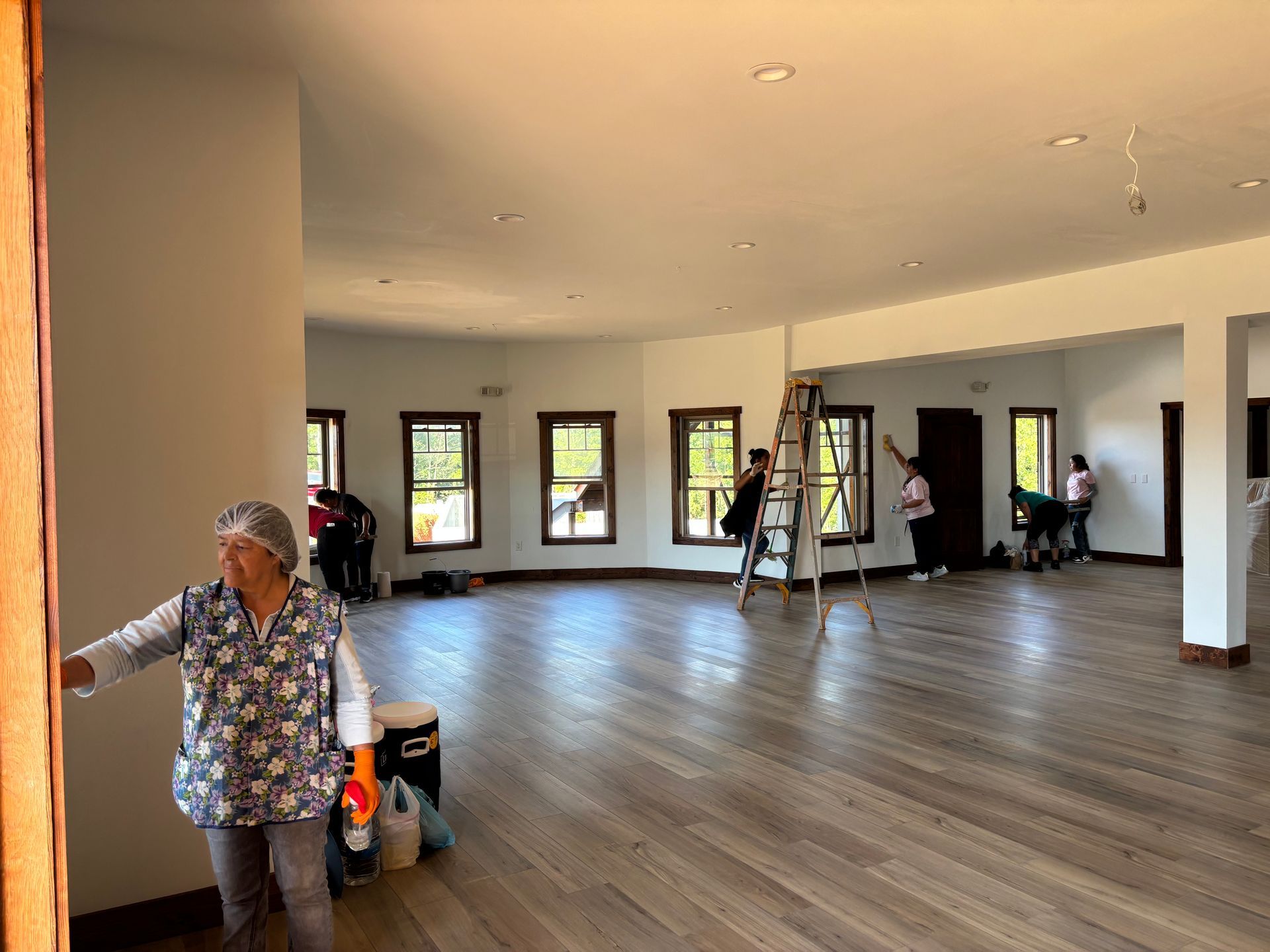 Large empty room with wood floors and windows, workers moving equipment during renovation