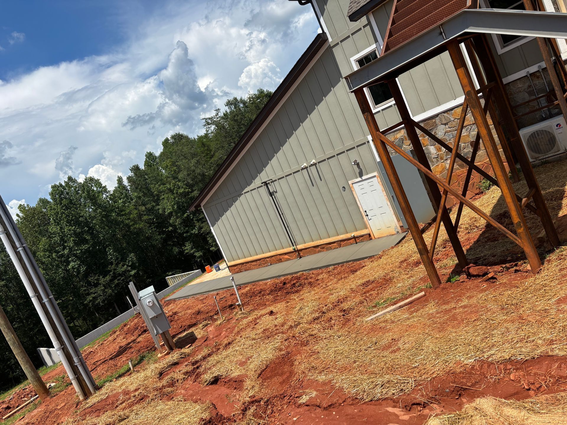 Tilted view of a rural metal building and wooden tower on red dirt under a cloudy sky