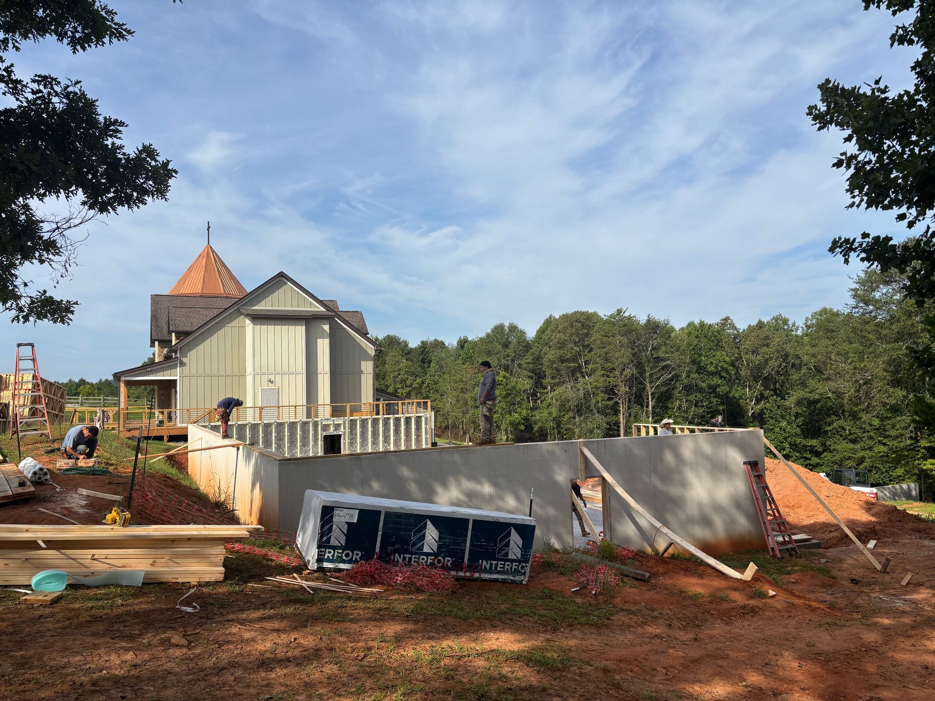 House under construction with concrete walls, workers, and orange tarps on a wooded hillside