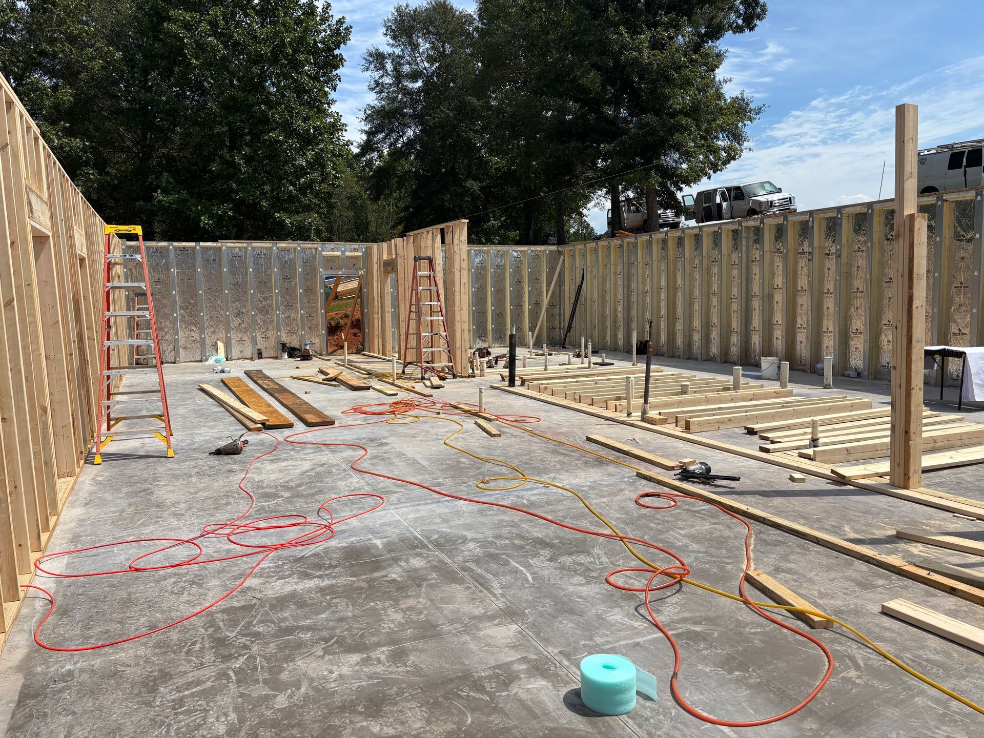 Construction site with wooden foundation walls, ladders, lumber, and cables on a concrete slab