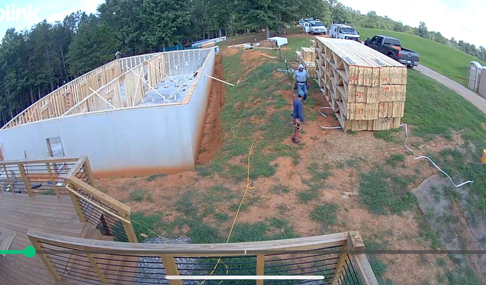 Construction site with concrete foundation walls, lumber stacks, and two workers on a dirt lot near trees