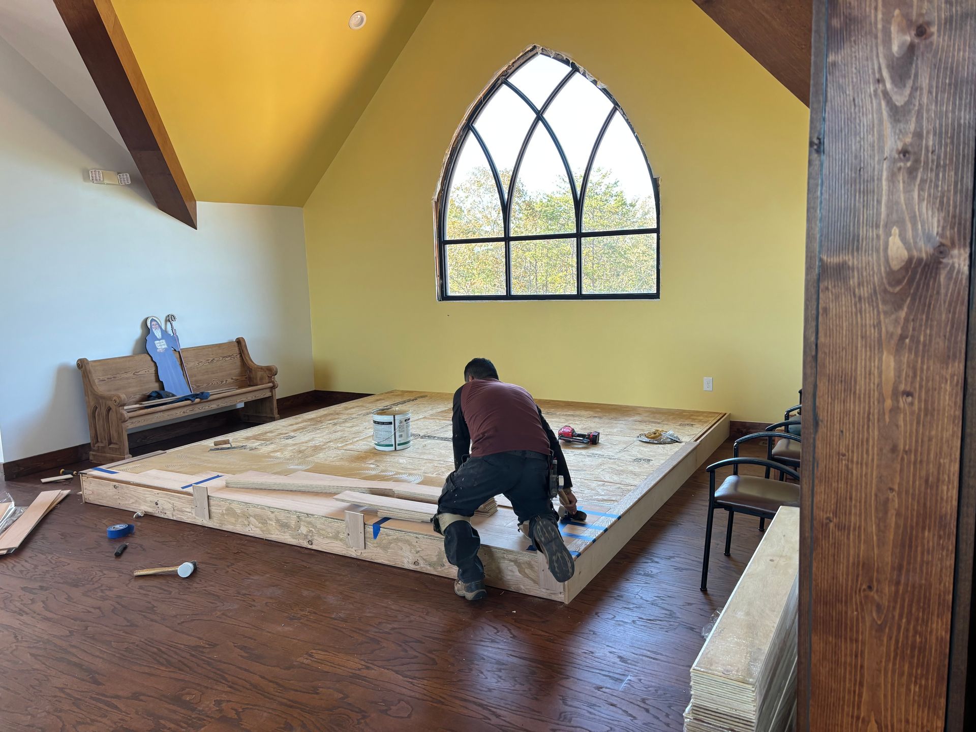 Person working on a raised wooden platform in a yellow room with a large arched window.