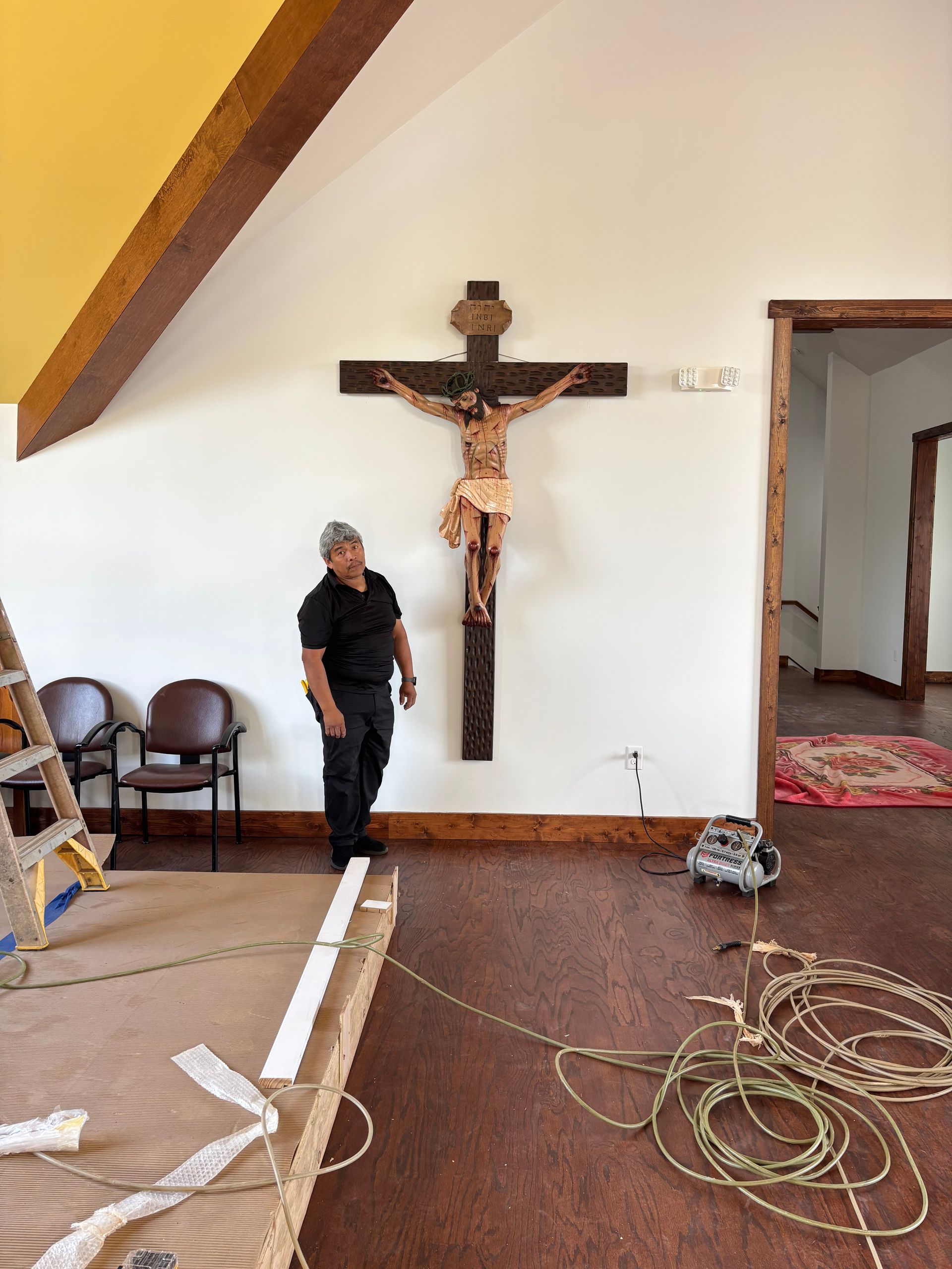 Man standing in a bare room beside a large crucifix mounted on a white wall, with construction debris on the floor.