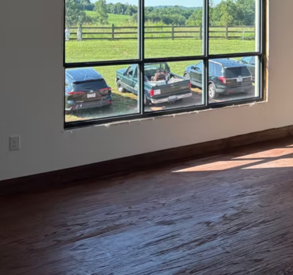 Empty room with wood floor and large windows overlooking parked cars and a green field