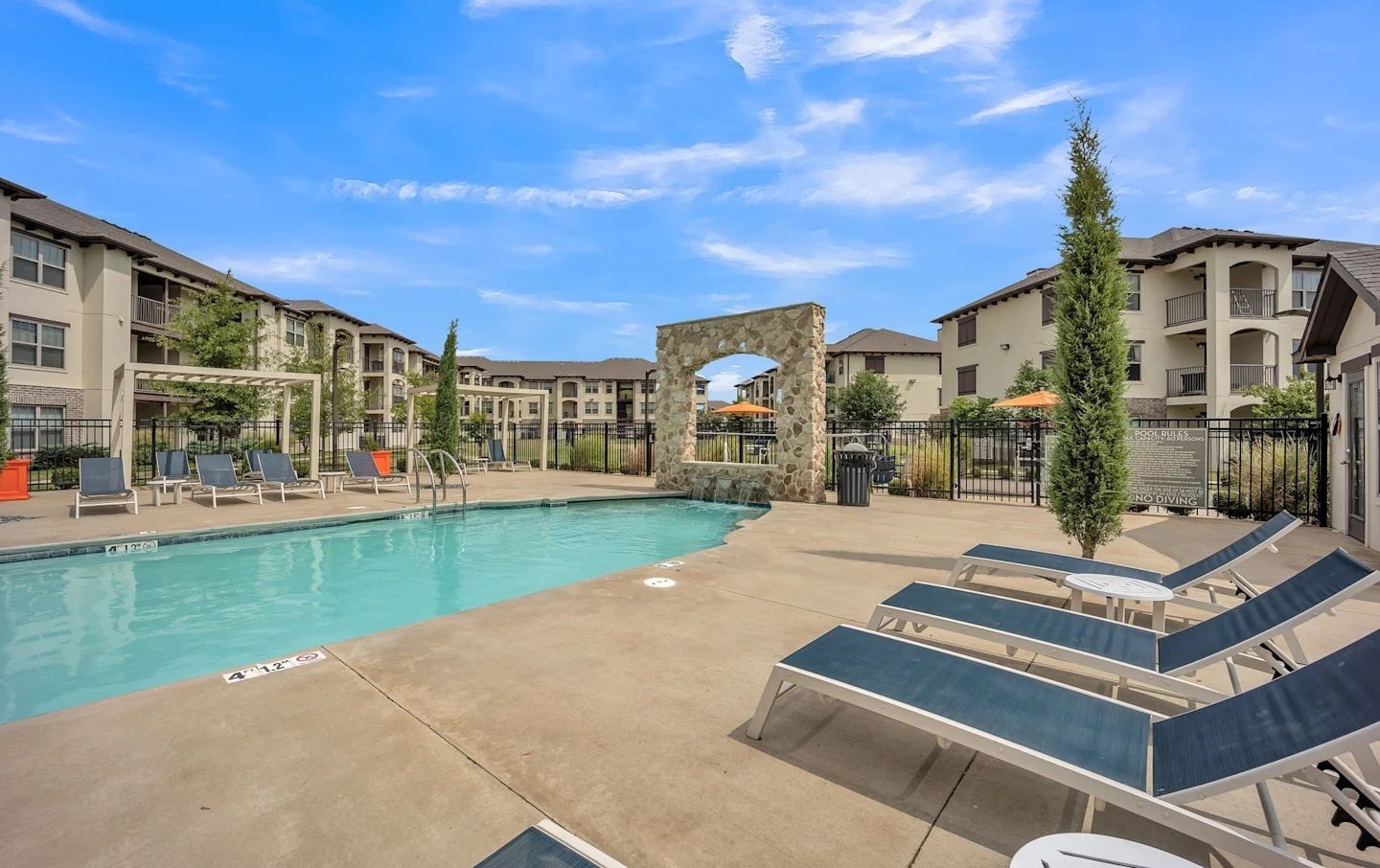 Swimming pool with lounge chairs, landscaping, and apartment buildings on a sunny day.
