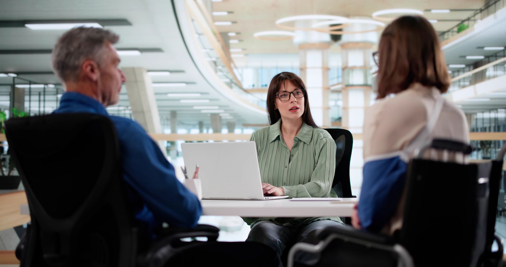 Three people in a bright office meeting, seated around a table and facing a woman speaking.