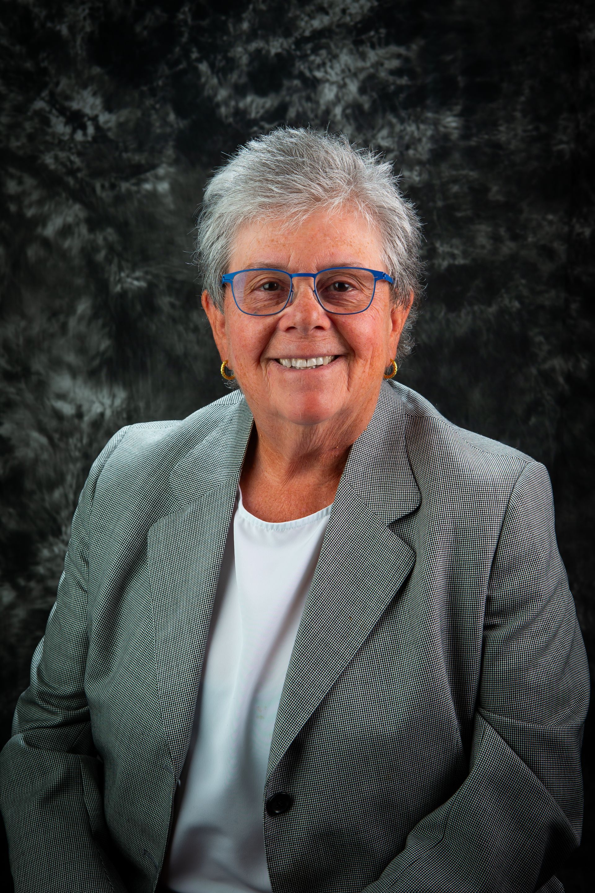 Person smiling in a gray checkered blazer and blue glasses against a dark studio backdrop