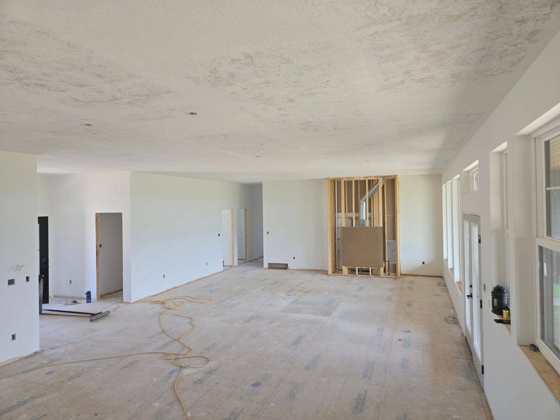 Interior view of a room under construction: white walls and ceiling, wood framing, unfinished floor, windows on the right.