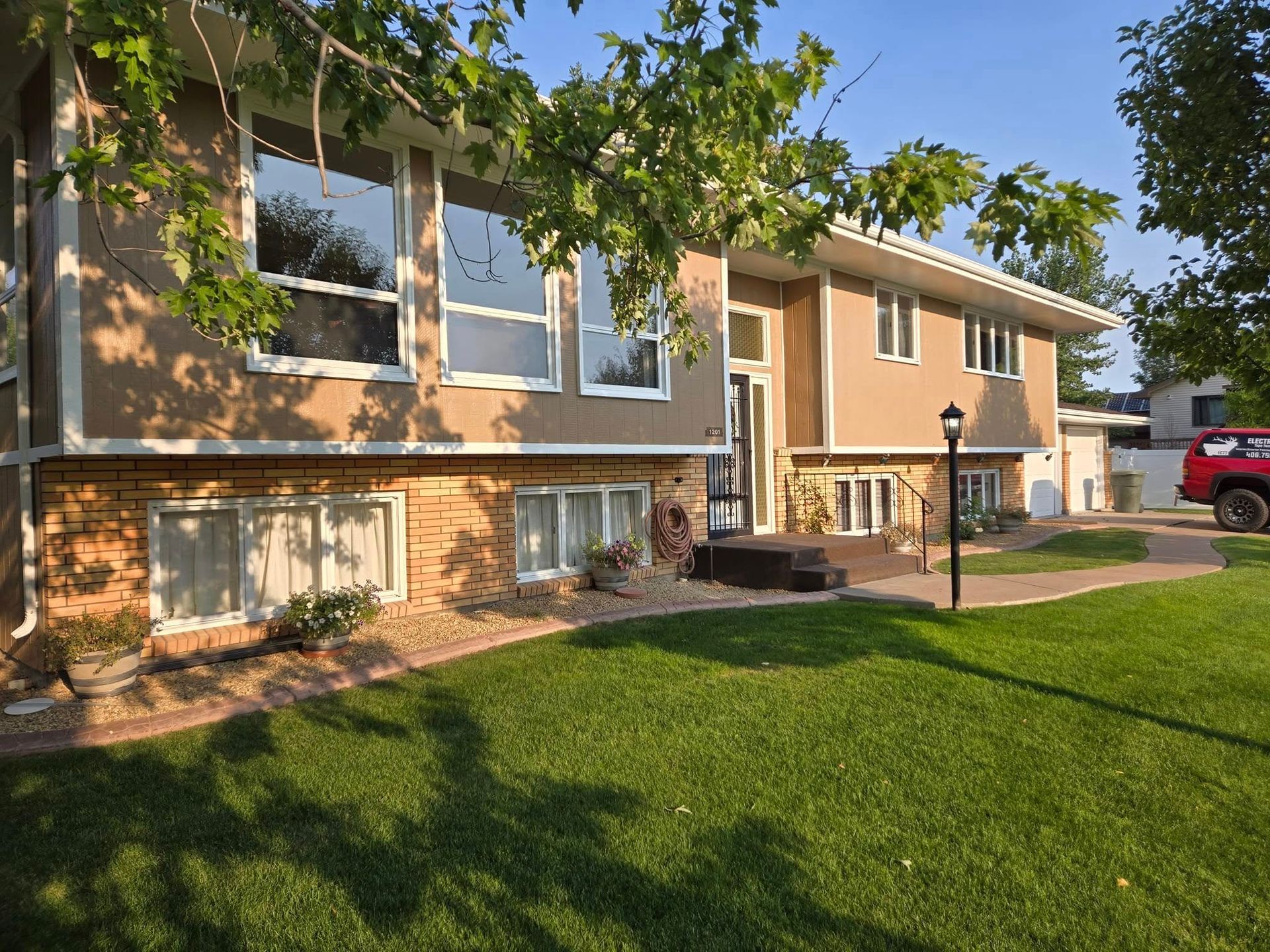 Two-story house with a brick base, tan siding, large windows, and a well-manicured lawn.
