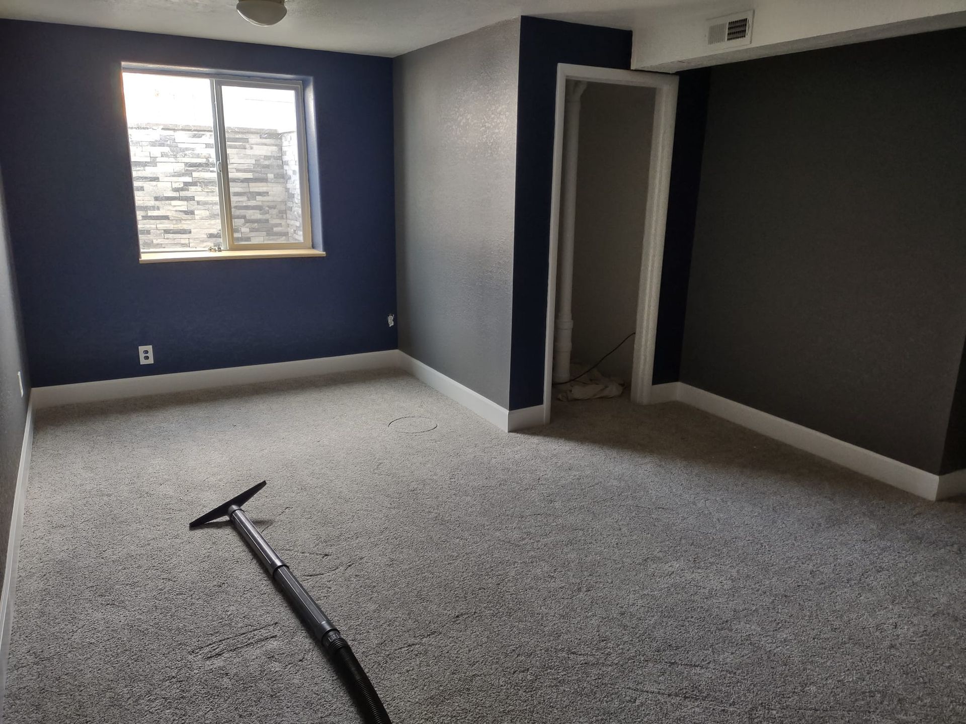 Empty room with dark gray walls, white trim, and gray carpet; a vacuum cleaner lies on the floor.