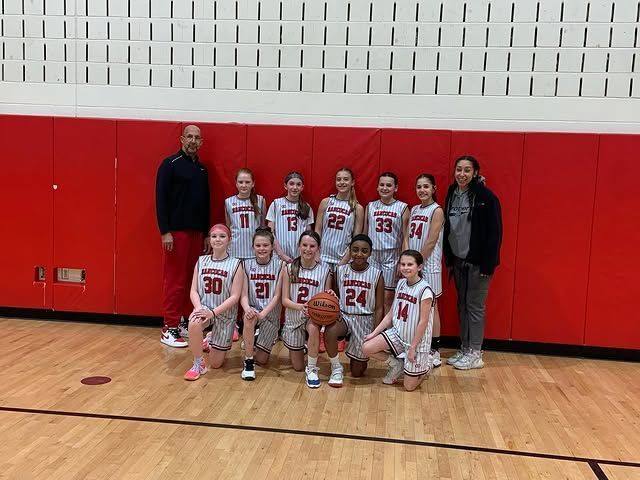 Youth basketball team posing in a gym, wearing white uniforms with red numbers in front of red wall padding
