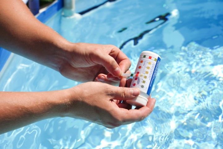 Hands testing a pool water test strip beside a blue swimming pool