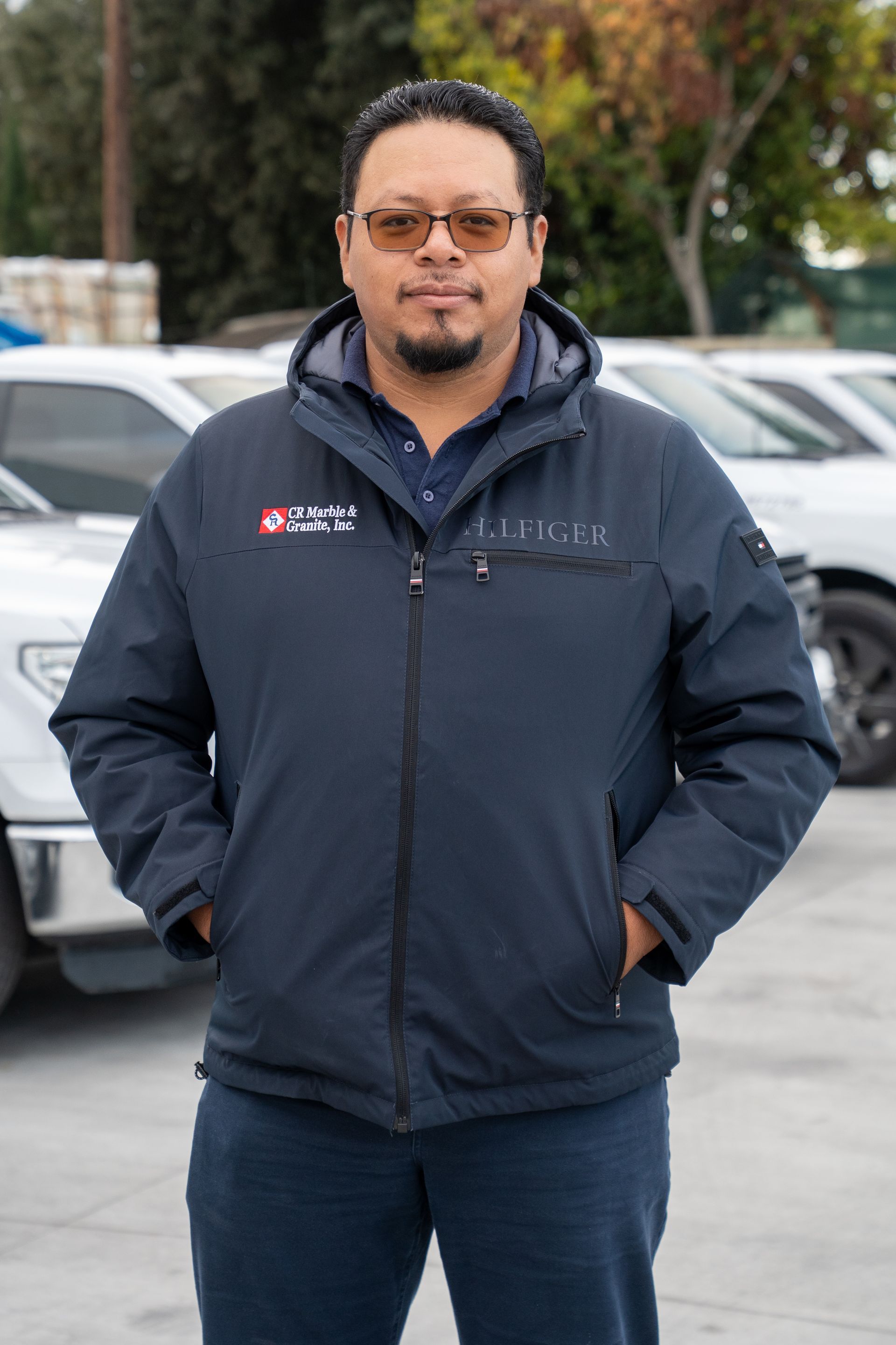 A man wearing a blue jacket and glasses is standing in front of a parking lot.