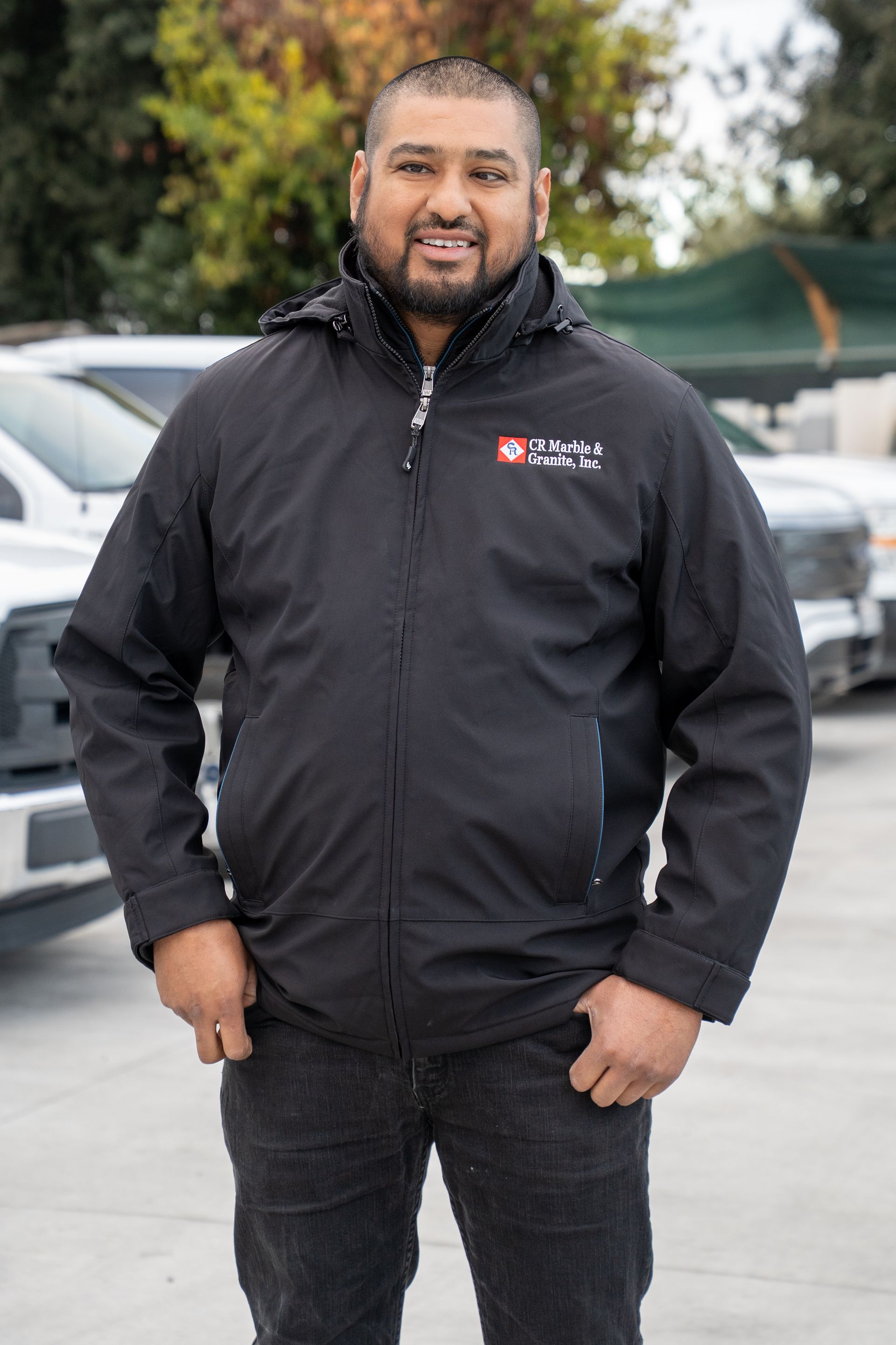 A man in a black jacket is standing in front of a row of cars.