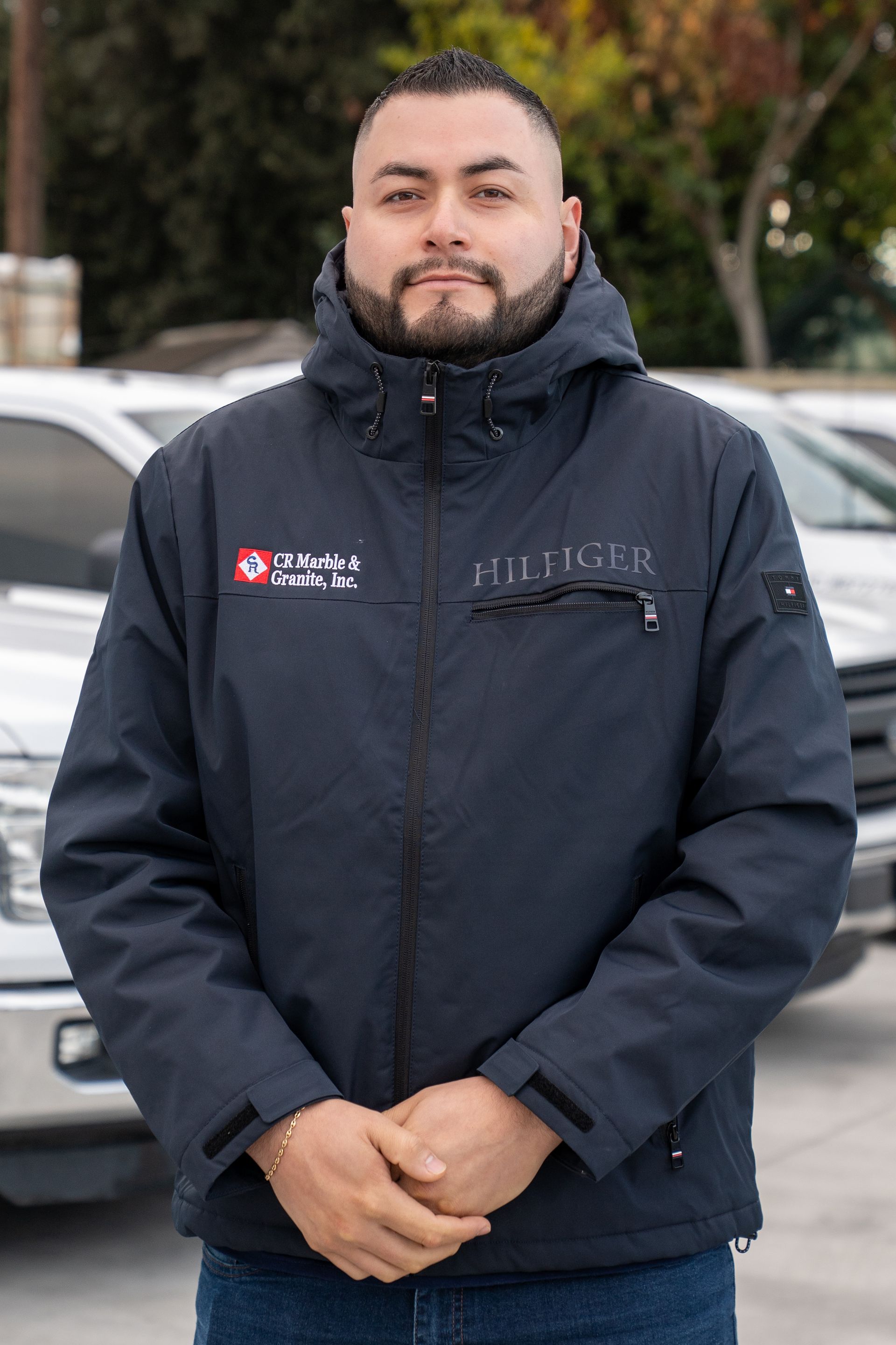 A man with a beard wearing a black jacket is standing in front of a row of cars.