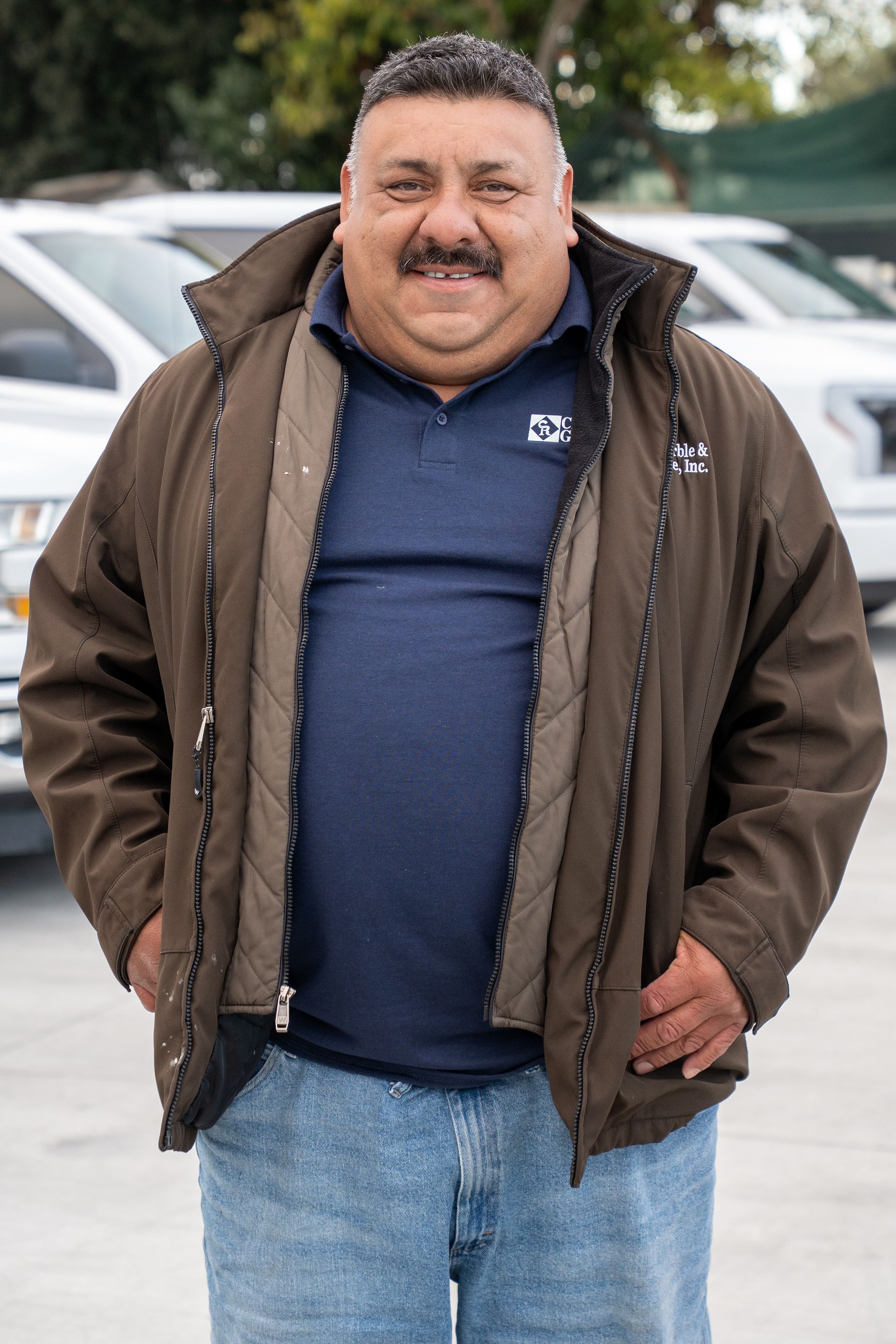 A man wearing a brown jacket and a blue shirt is standing in a parking lot.