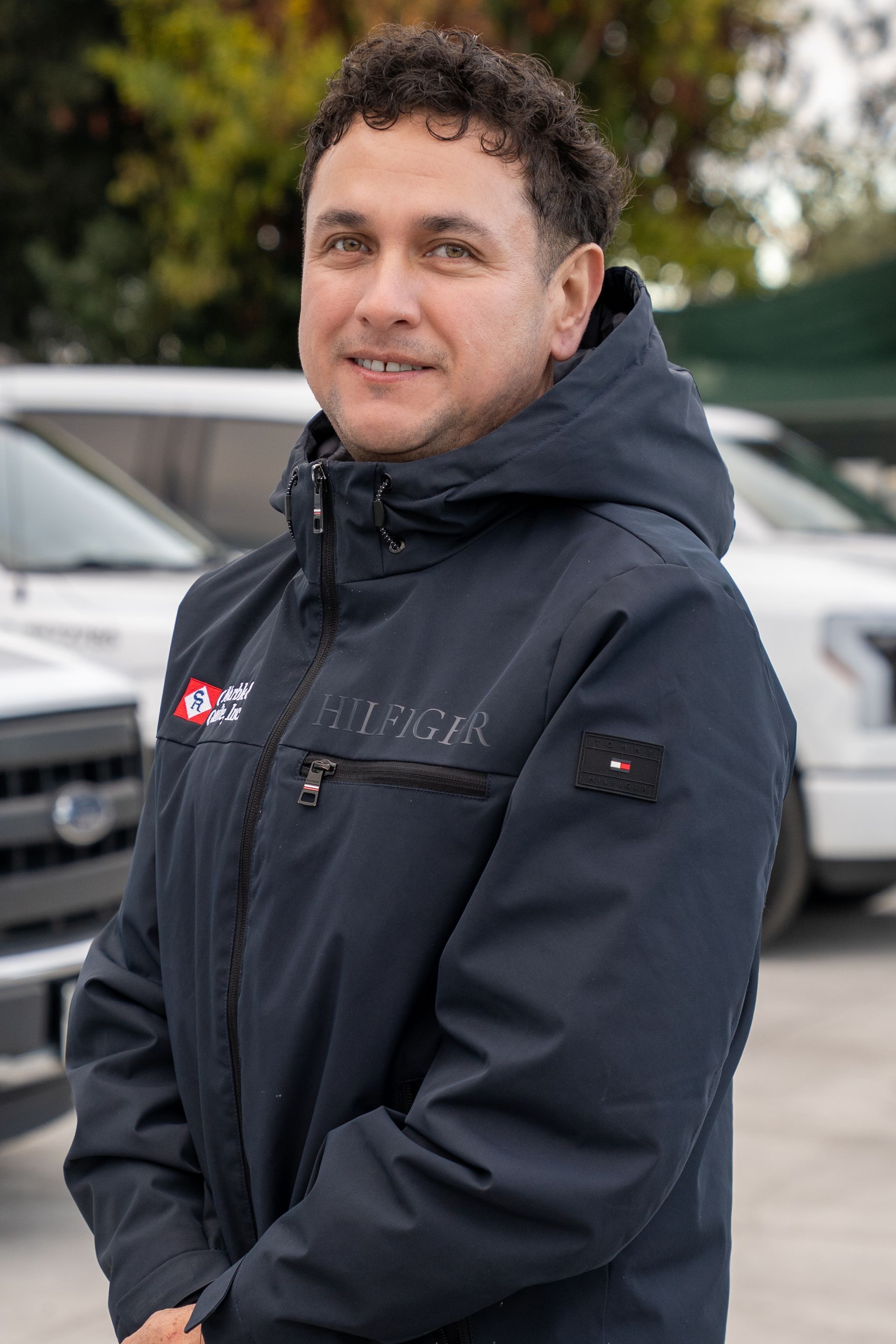 A man in a black jacket is standing in front of a row of cars.