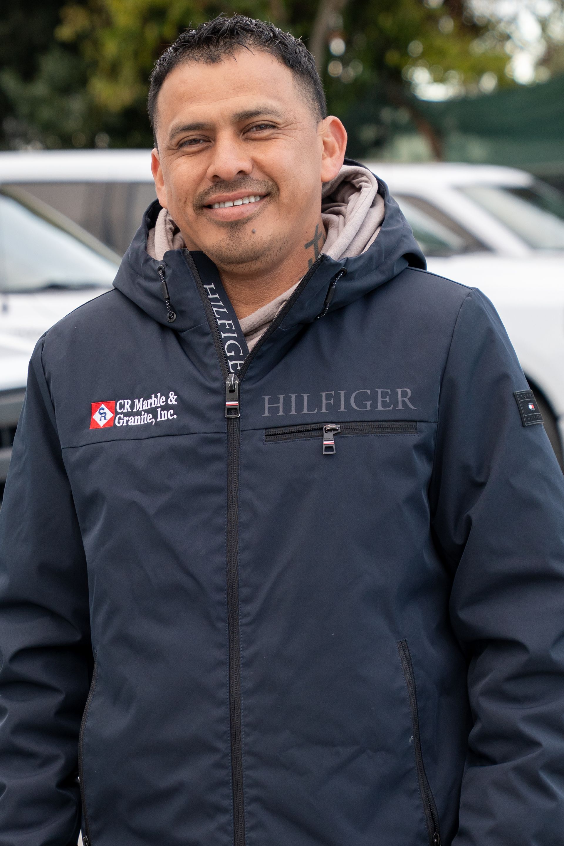 A man wearing a hilfiger jacket is standing in front of a row of cars.