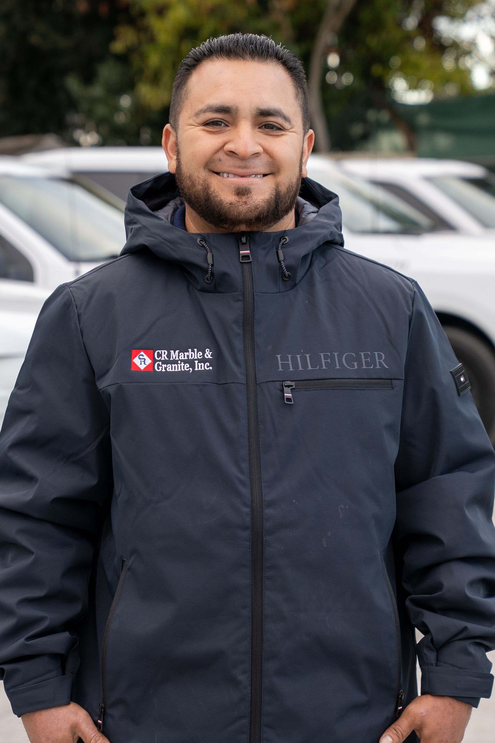 A man with a beard wearing a tommy hilfiger jacket is standing in front of a parking lot.