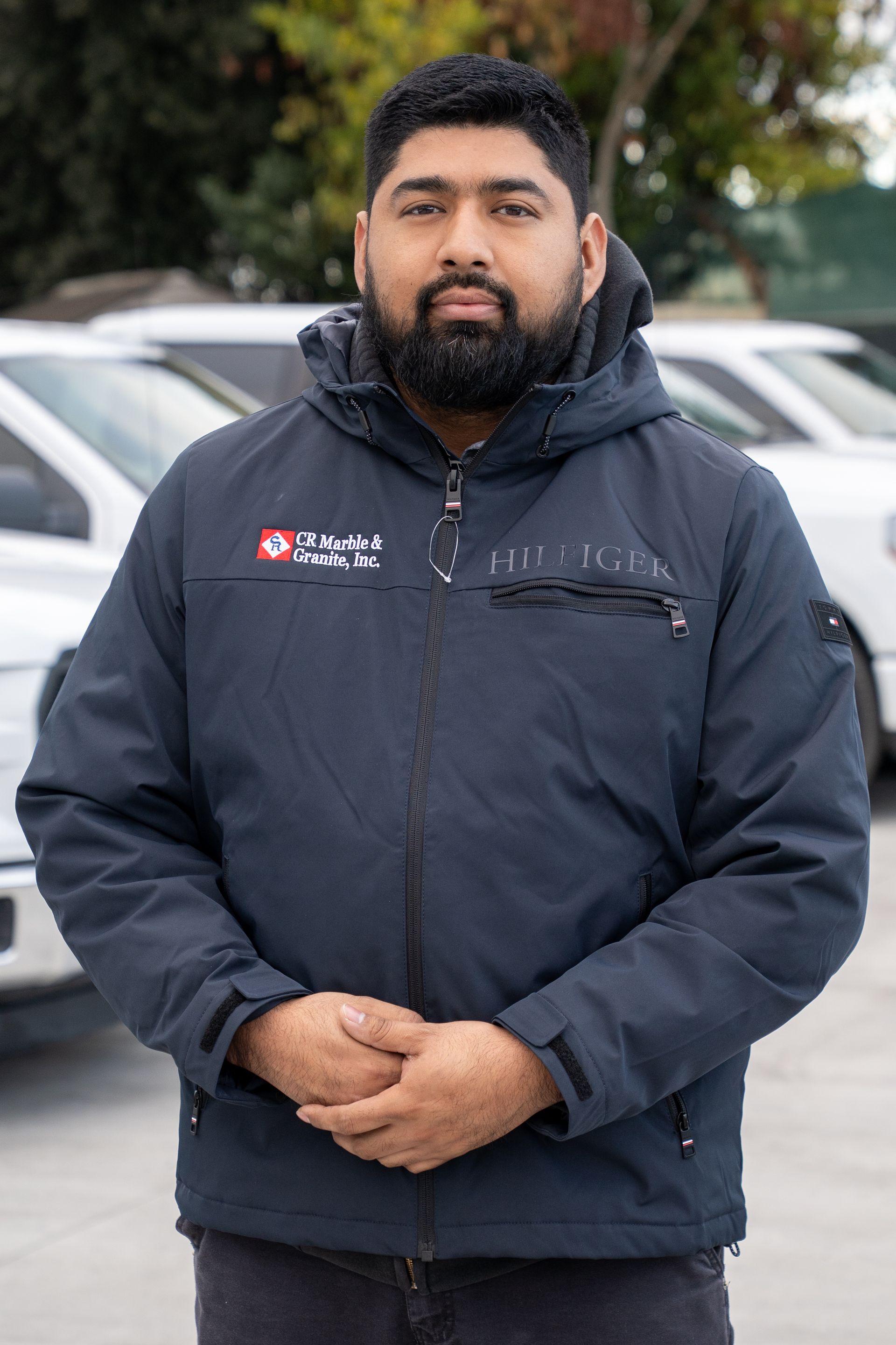 A man with a beard is standing in front of a row of cars in a parking lot.