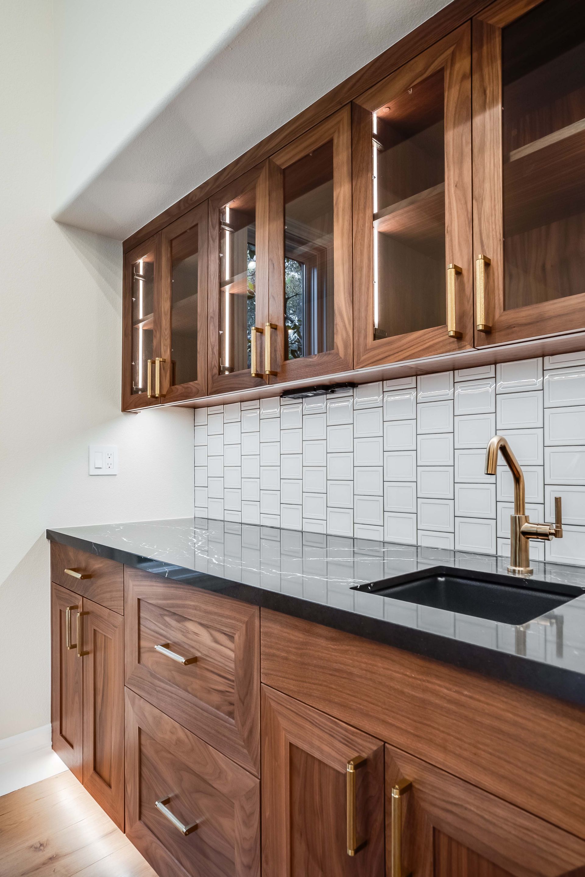 Wooden cabinets in a kitchen, with a black countertop, white backsplash, and gold fixtures.