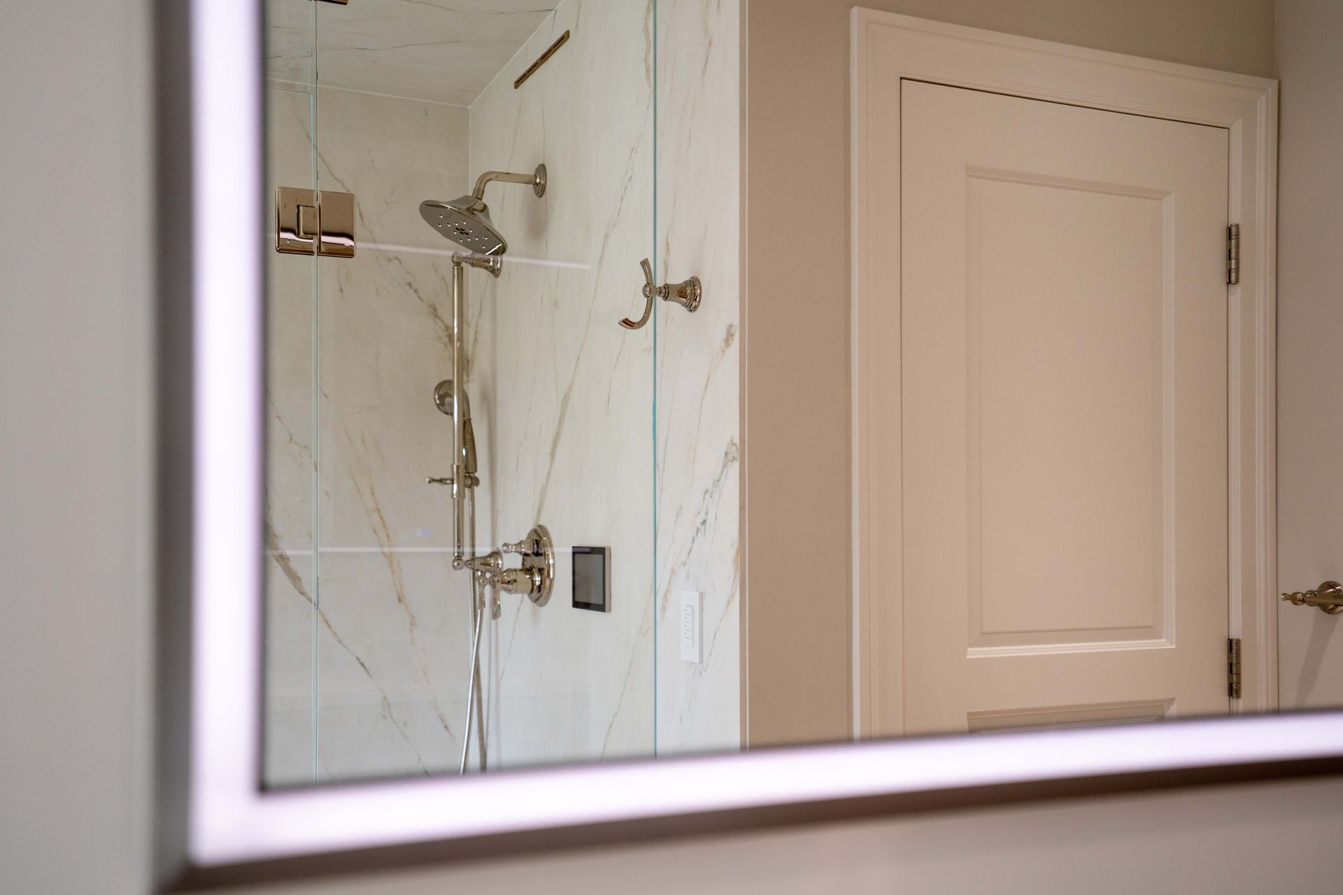 Mirror reflecting a white and gold bathroom. Shower with glass doors and a white door are visible.