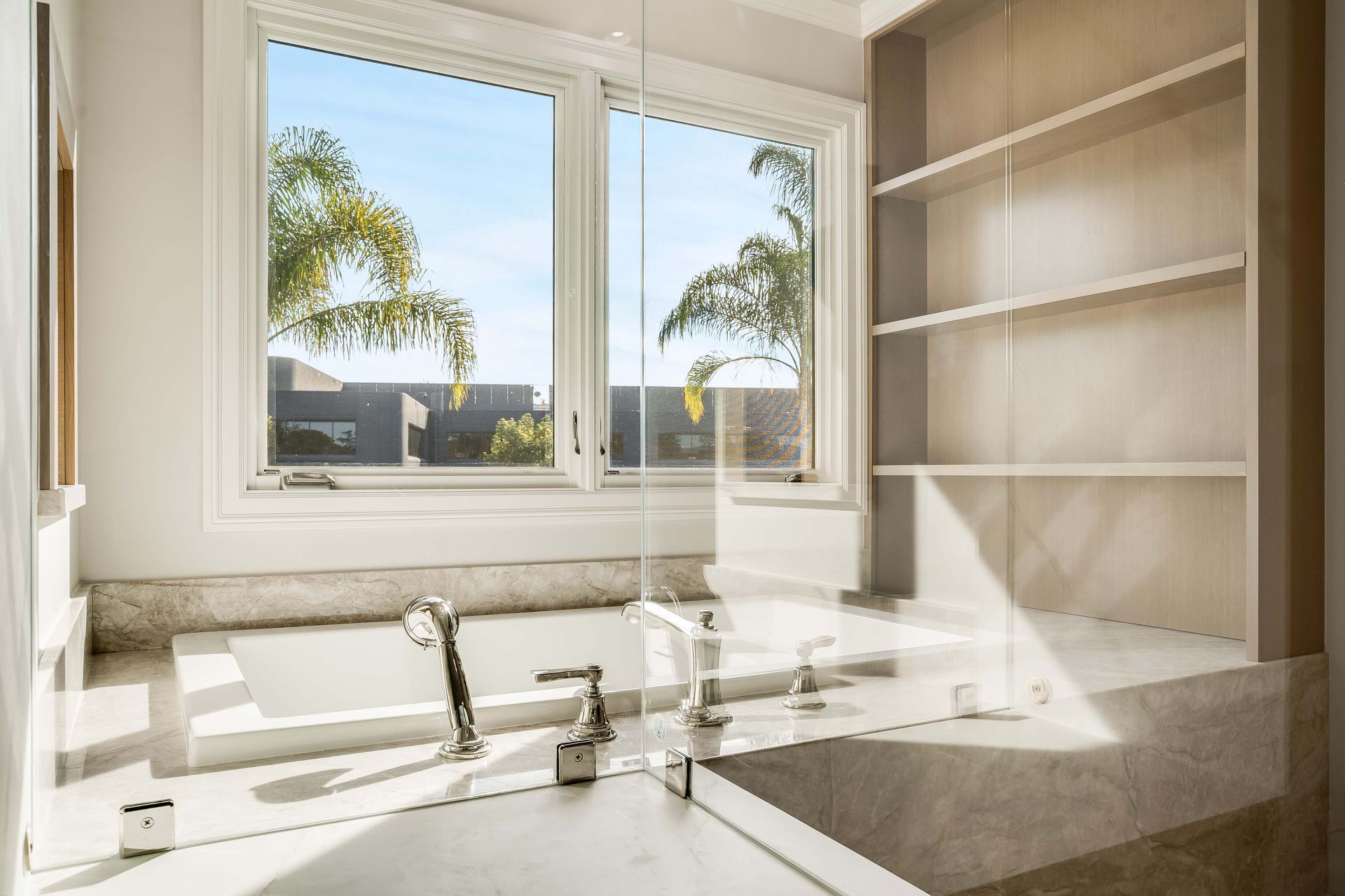 Bathroom with a built-in tub, large window overlooking palm trees, and built-in shelving.
