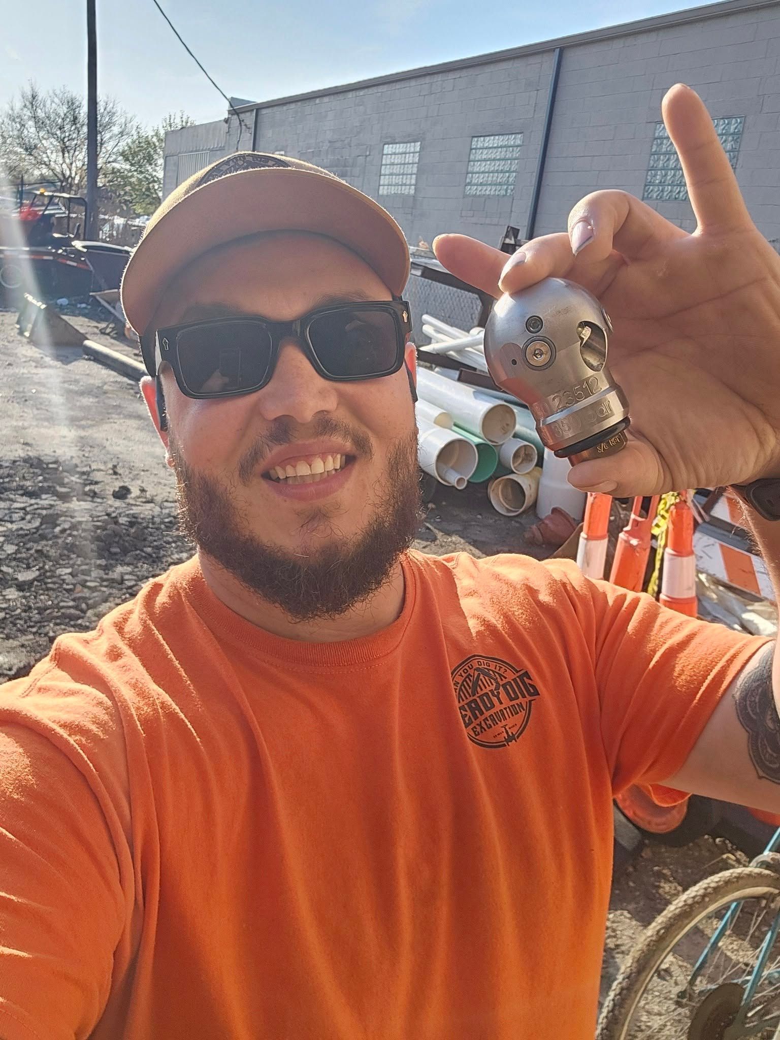 Smiling man in sunglasses and orange shirt holding up a small metallic object at an outdoor work site
