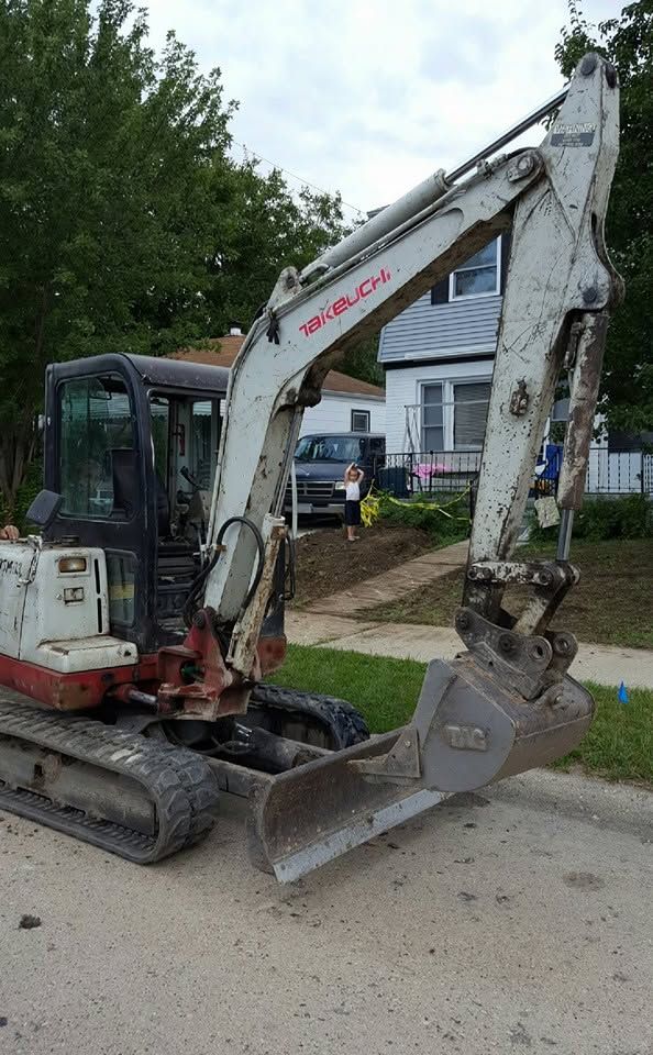 White excavator on a street with bucket lowered beside a residential house and trees