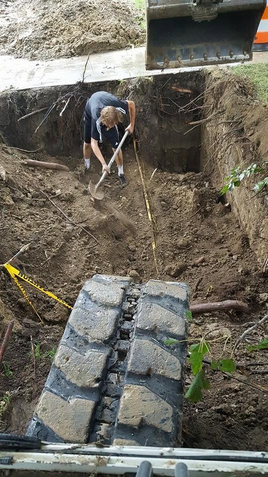 Worker digging in a muddy trench beside a concrete structure, with stone blocks in the foreground.
