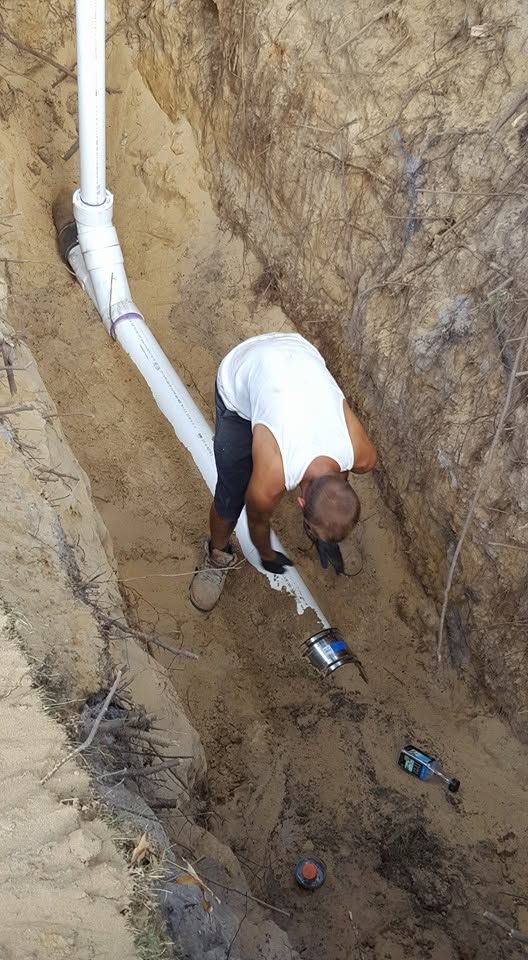 Worker bending in a dirt trench beside white PVC pipes, using a tool near an underground pipe joint.