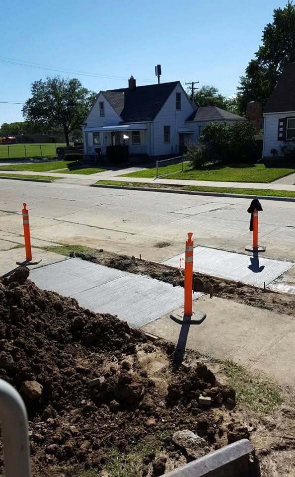 Sidewalk construction with open trench, orange markers, and a house across the street on a sunny day