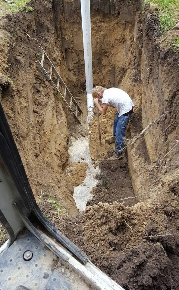 Worker digging a deep trench beside a white pipe in muddy soil
