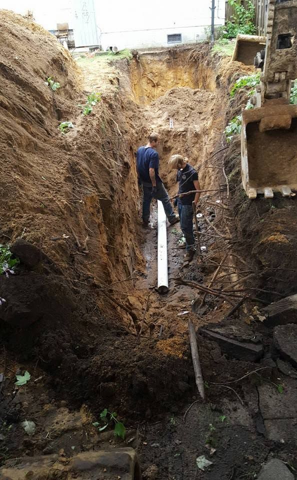 Two workers inspecting a white pipe in a deep, muddy excavation site.