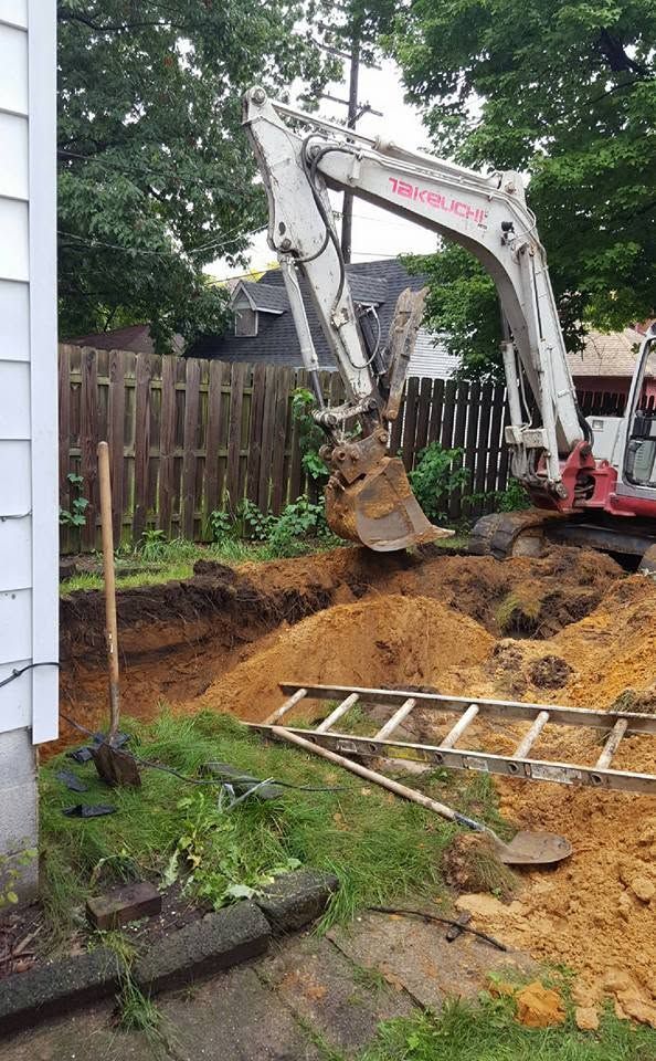 Excavator digging a large backyard hole beside a wooden fence, with a ladder on the ground.