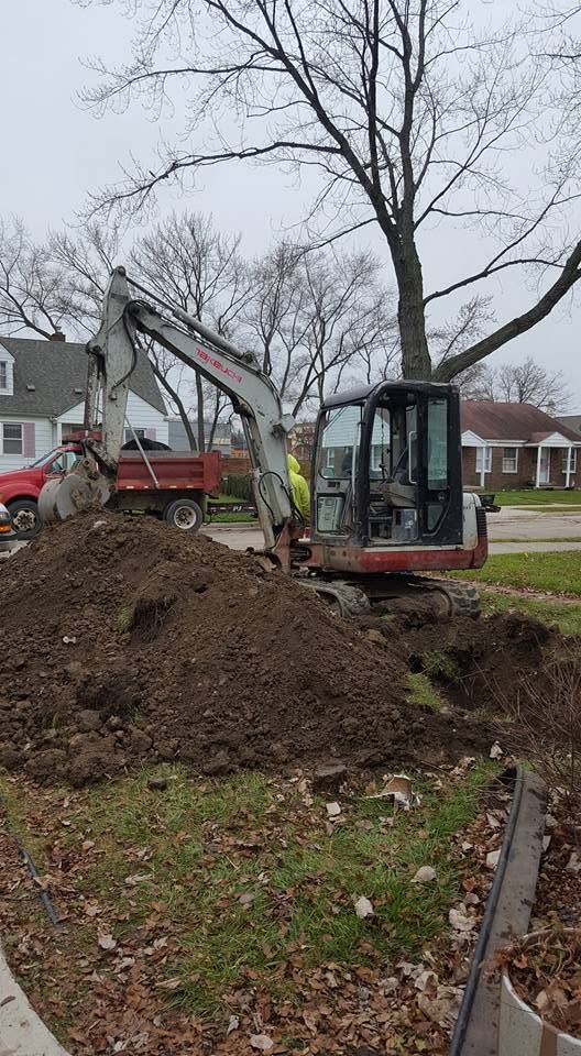Excavator digging a large dirt pile in a suburban yard with bare trees and houses nearby