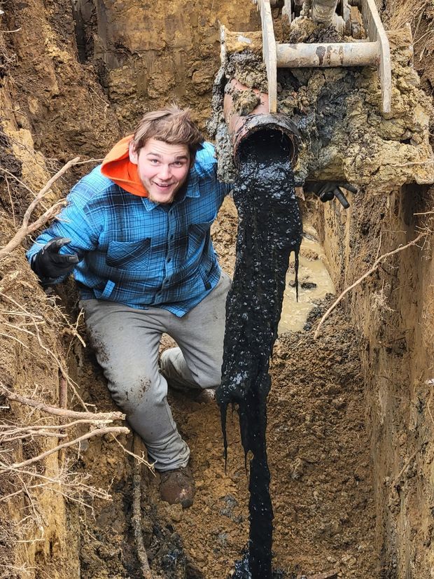 Person in blue jacket standing in a muddy trench beside a backhoe bucket and pipe.