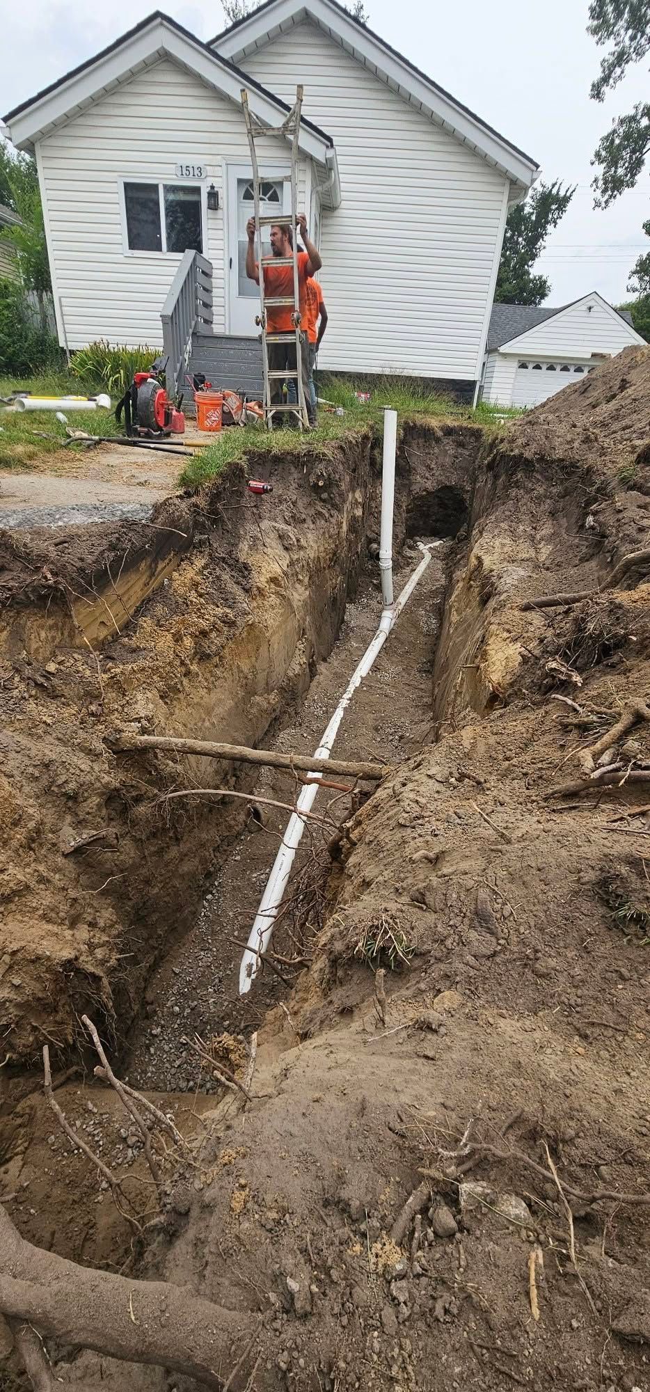 Worker in orange vest surveys a deep trench with a white pipe beside a white house.